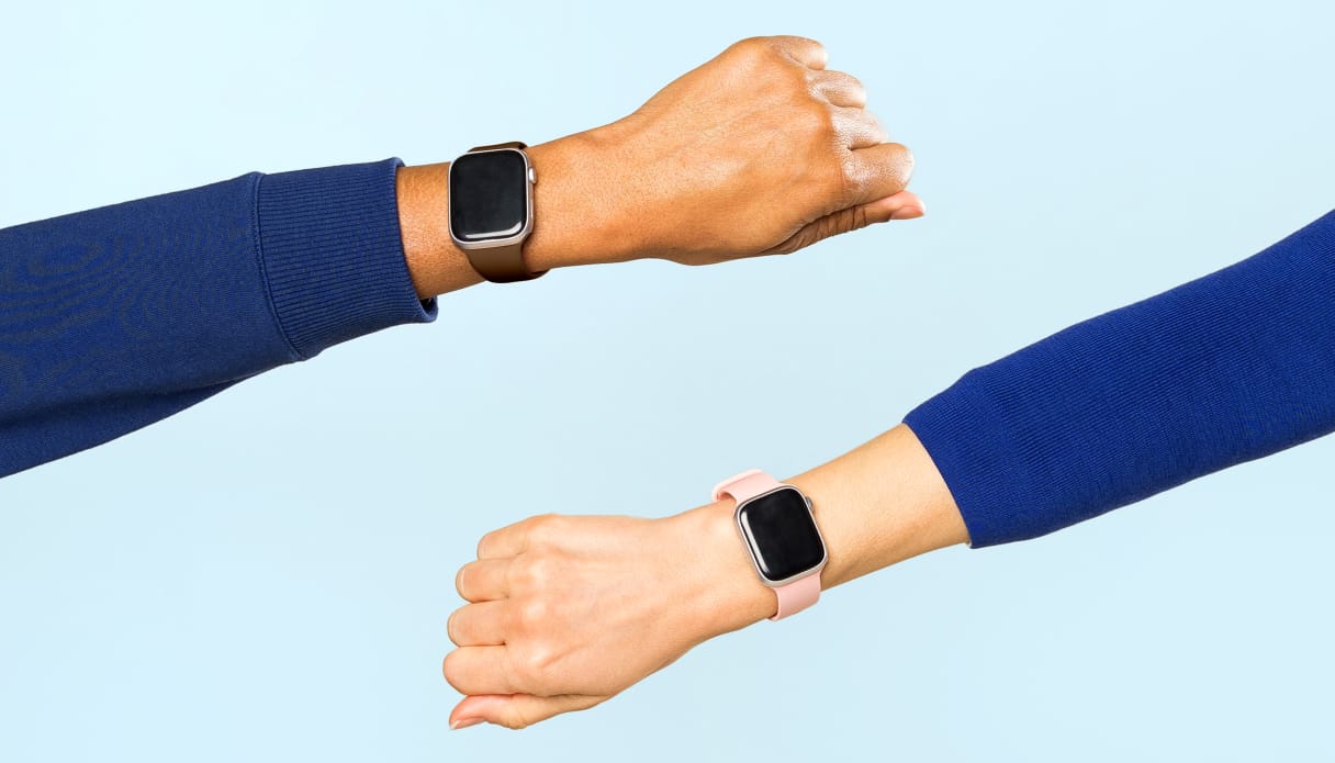 Zwei Unterarme kommen von beiden Seiten ins Bild, beide mit einer Smartwatch am Handgelenk, einem blauen Pullover und hellblauem Hintergrund.
