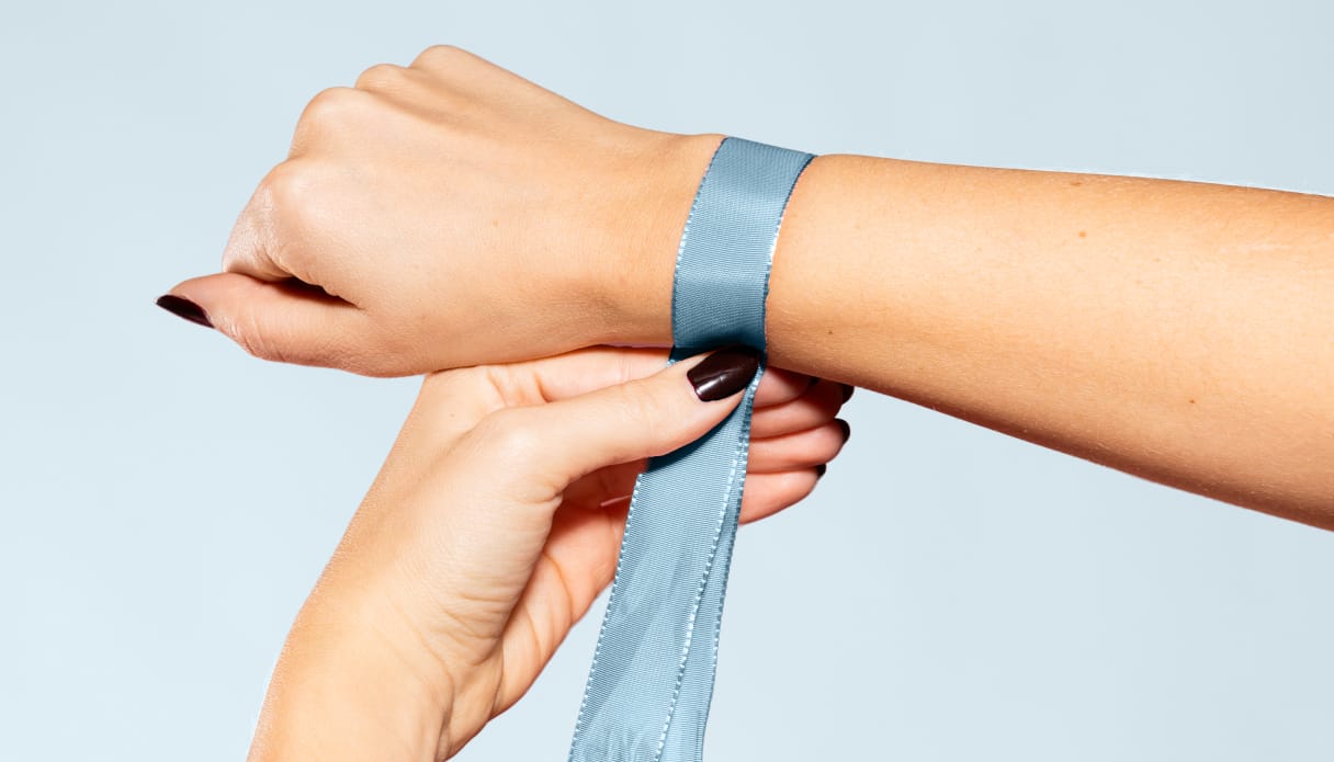 Eine Hand mit einem blauen Band um das Handgelenk, mit dem der Handgelenkumfang gemessen wird; der Hintergrund ist hellblau.