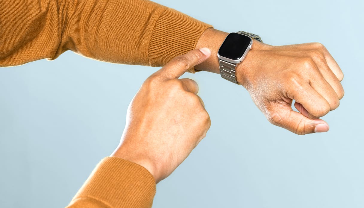 Zu sehen ist ein Arm mit einer Smartwatch am Handgelenk vor hellblauem Hintergrund. Der Pullover ist gelb und die Smartwatch wird mit der anderen Hand bedient.