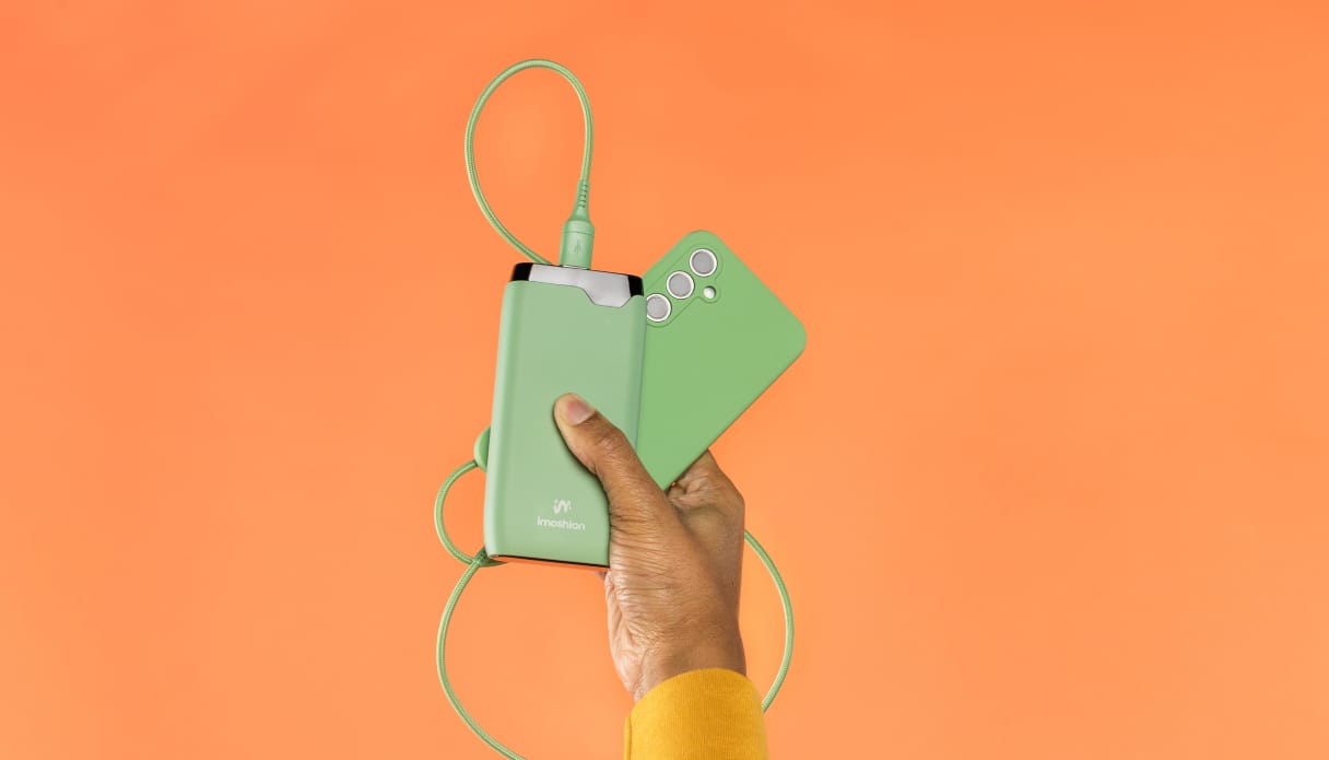 Eine Hand mit einem Stück gelbem Ärmel ragt von unten hervor und hält ein Handy mit grüner Hülle, ein grünes Ladekabel und eine grüne imoshion Powerbank fest. Der Hintergrund ist orange.