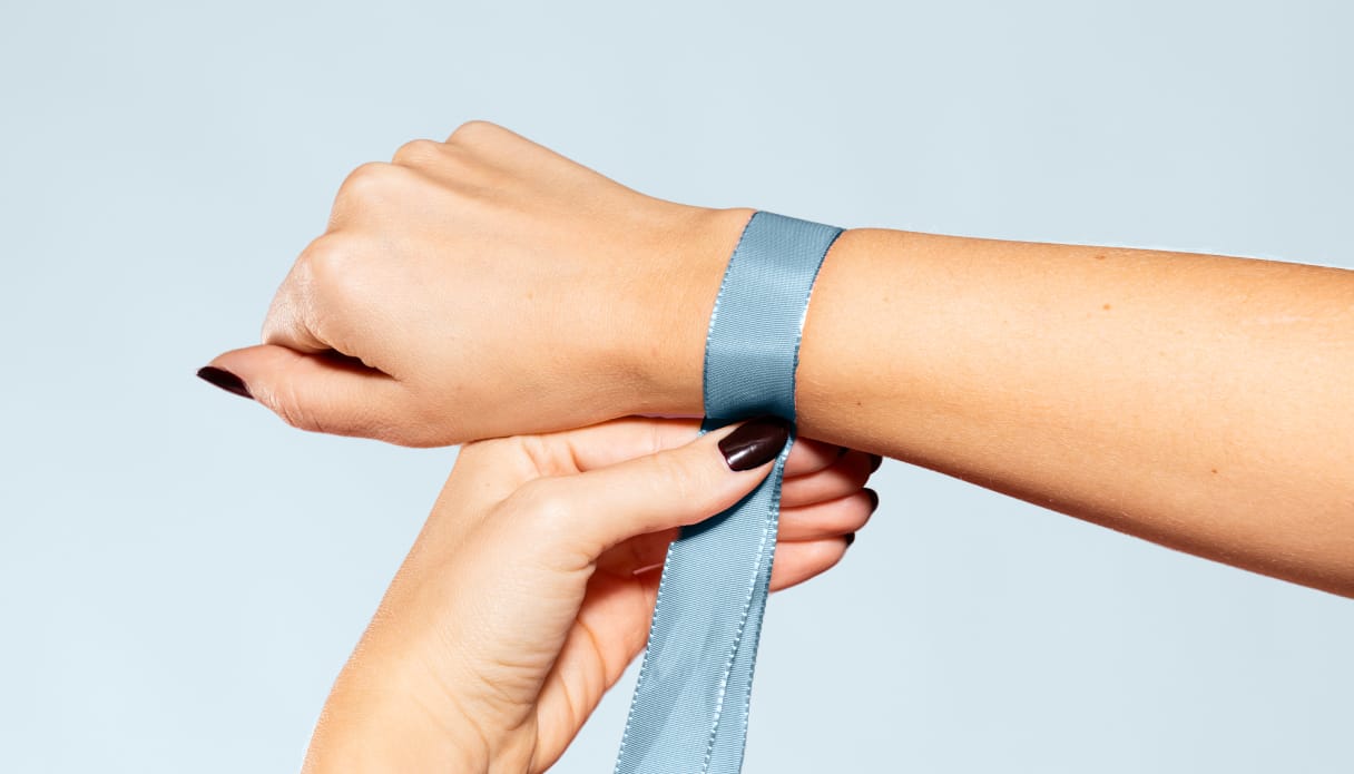 Eine Hand mit einem blauen Maßband um das Handgelenk, mit dem der Handgelenkumfang gemessen wird; der Hintergrund ist hellblau.