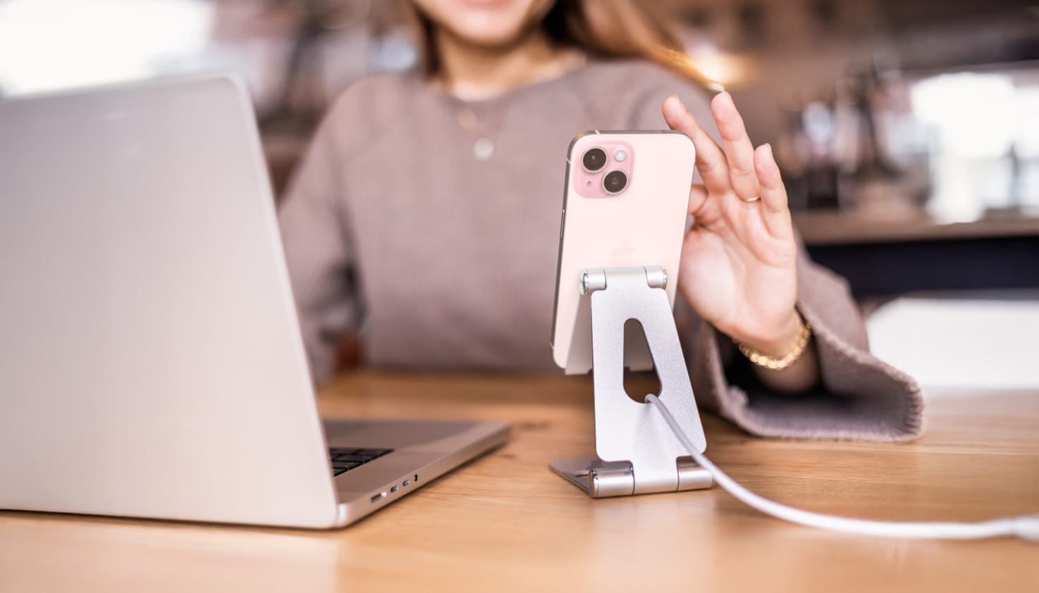 een vrouw bedient haar telefoon die in een telefoonstandaard staat terwijl hij wordt opgeladen. Op de tafel staat ook haar laptop.