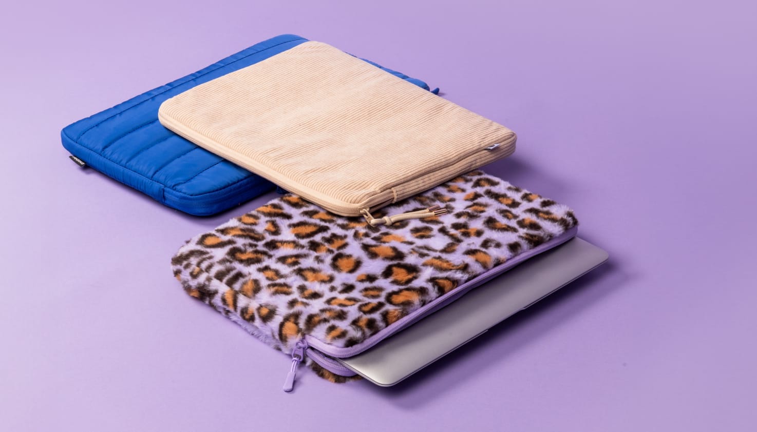 Eine kobaltblaue Laptop-Hülle, eine beige Cord-Hülle und eine Laptop-Hülle mit violettem Leopardenmuster liegen nebeneinander.