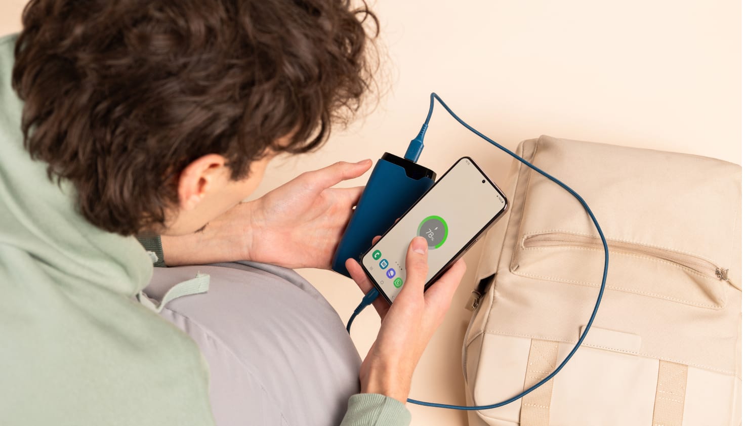 Du siehst eine Person mit braunen Haaren, die auf ein Smartphone schaut, das über ein blaues Kabel an eine blaue Powerbank angeschlossen ist. Im beigen Hintergrund liegt ein sandfarbener Rucksack.