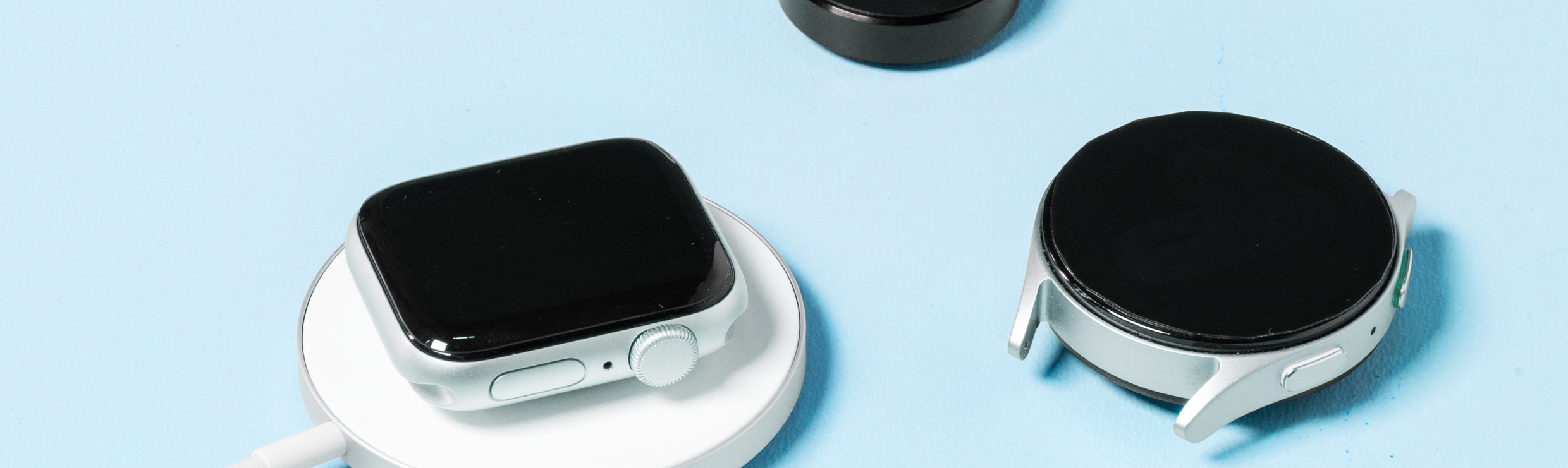 Eine Apple Watch auf einem kabellosen Ladegerät, daneben liegt eine Samsung Watch und ein separates Smartwatch-Ladegerät.